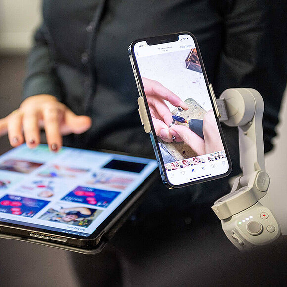Ein Smartphone auf einer Halterung. Im Hintergrund ist unscharf eine Frau zu sehen, die auf einem Tablet tippt.